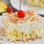 close up view of square piece of pina colada poke cake topped with frosting and toasted coconut on a white plate with a cherry on top - 22