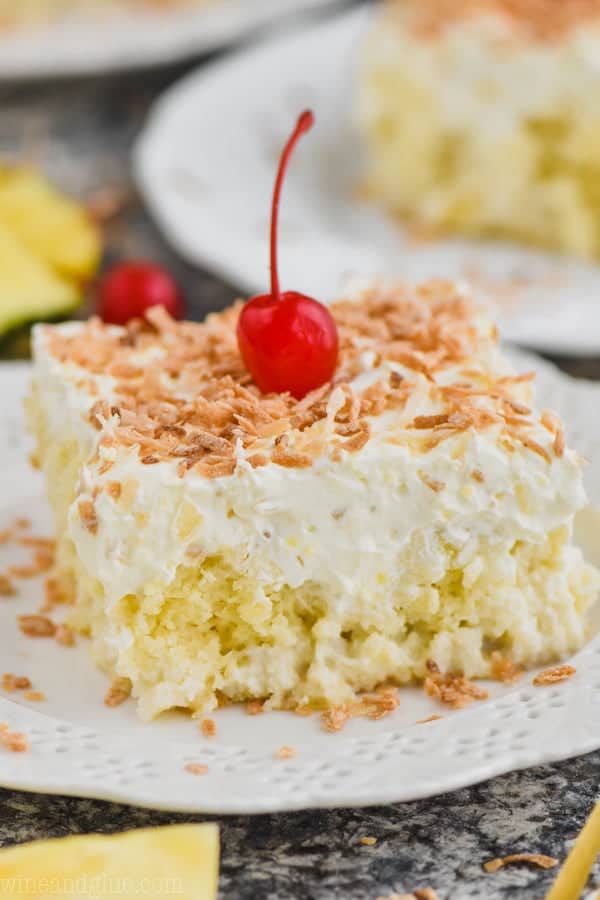 close up view of square piece of pina colada poke cake topped with frosting and toasted coconut on a white plate with a cherry on top