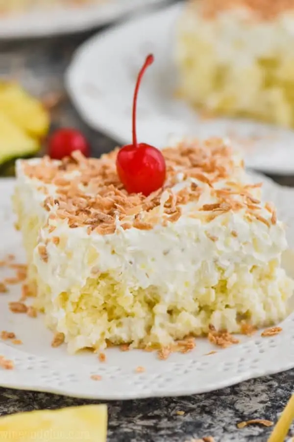 close up view of square piece of pina colada poke cake topped with frosting and toasted coconut on a white plate with a cherry on top
