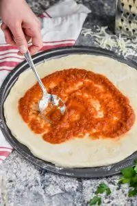 woman using a metal spoon to spread pizza sauce onto a circle pizza pan with dough on it