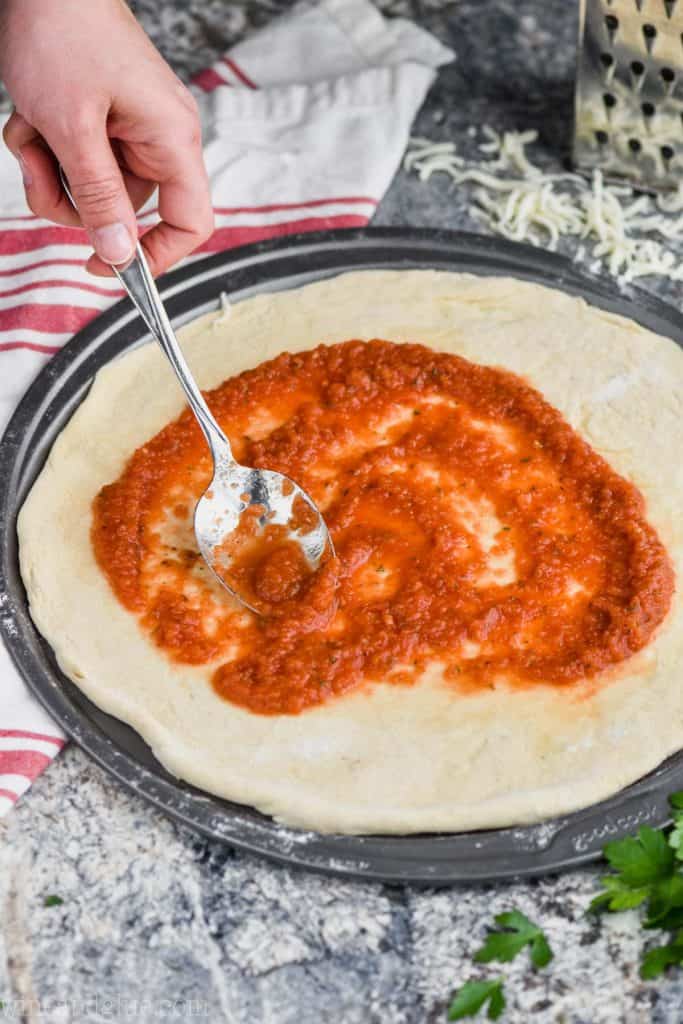 woman using a metal spoon to spread pizza sauce onto a circle pizza pan with dough on it