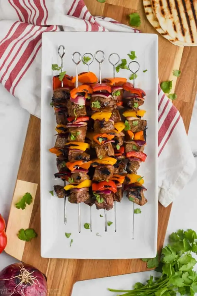 overhead view of beef kabobs on a white plate on a cutting board with grilled tortilla shells to make steak fajitas