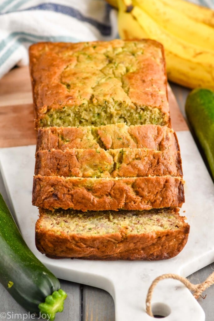 Overhead photo of a loaf of Banana Zucchini Bread that is partially sliced. Bananas and zucchini sit beside cutting board.