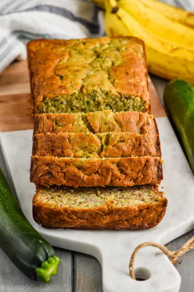 front view of zucchini banana bread recipe sliced on a marble and wood cutting board surrounded by two zucchini and a bunch of bananas on a gray wood board