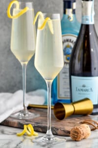 Photo of two French 75 cocktails. Bottles of gin and champagne for cocktail are behind the cocktails.