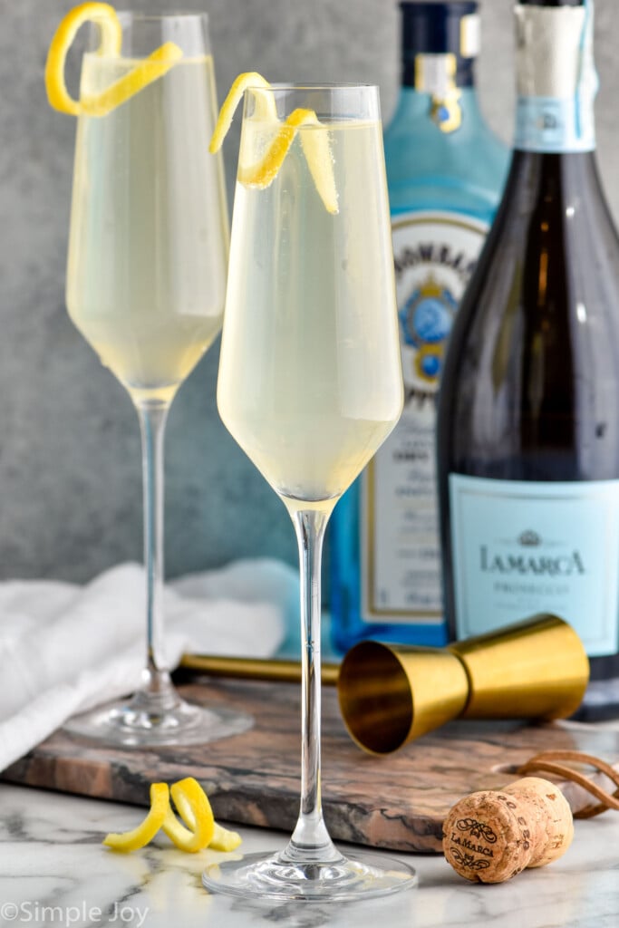 Photo of two French 75 cocktails. Bottles of gin and champagne for cocktail are behind the cocktails.