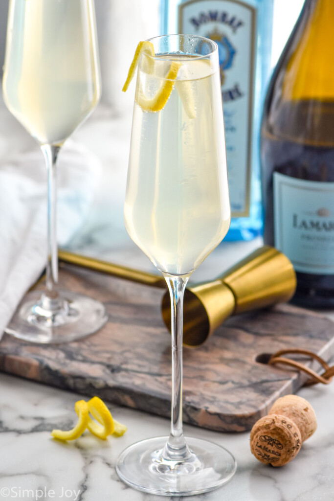 Close up photo of French 75 cocktail garnished with lemon peel. Bottles of gin and champagne are in the background along with a measuring cup.