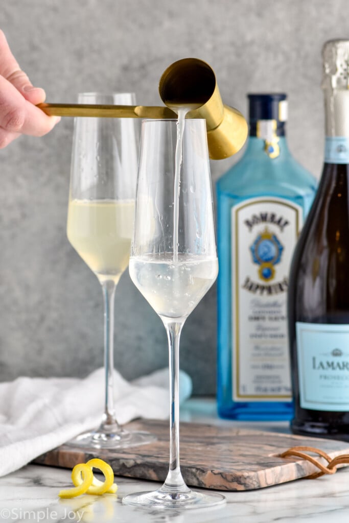 Photo of person's hand pouring ingredients into champagne flutes for French 75 cocktails. Bottles of gin and champagne are in the background.