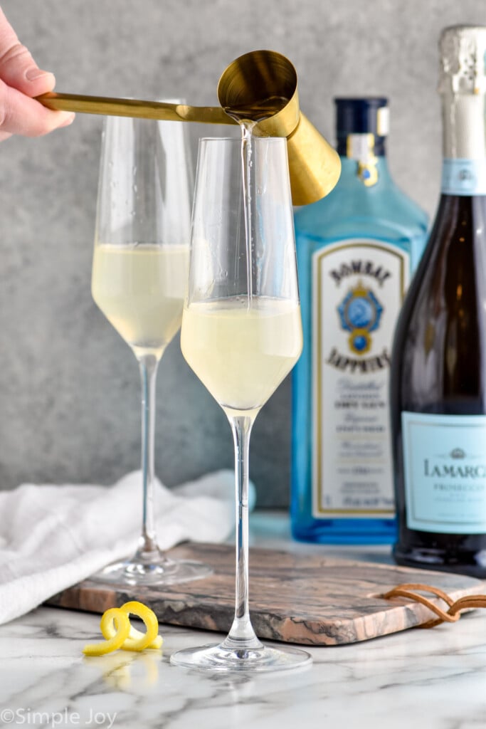 Photo of person's hand pouring ingredients into champagne flutes for French 75 cocktails. Bottles of gin and champagne are in the background.