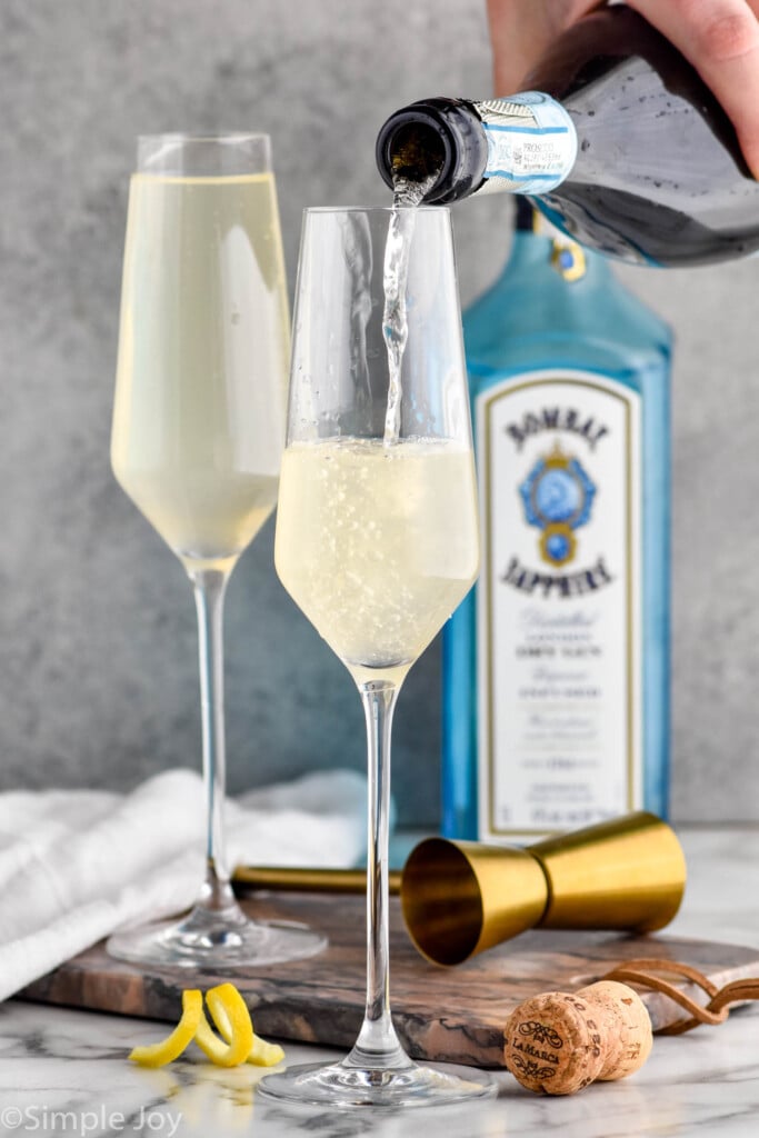 Photo of person's hand pouring champagne into flutes for French 75 cocktail. Bottle of gin is in the background.