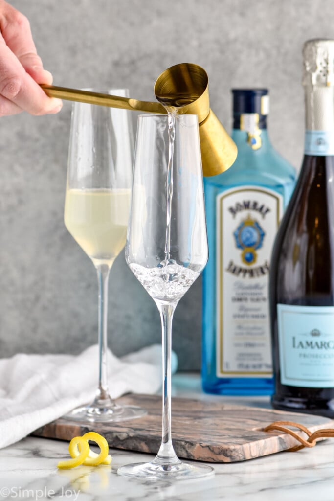 Photo of person's hand pouring ingredients into champagne flutes for French 75 cocktails. Bottles of gin and champagne are in the background.