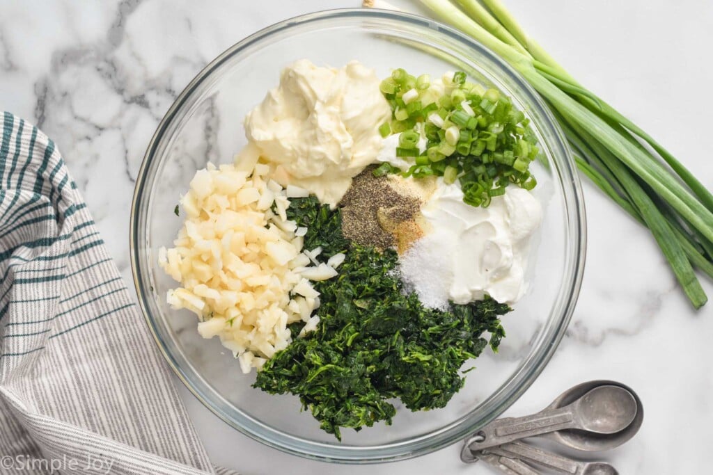 Overhead photo of ingredients in clear bowl for Spinach Dip Recipe. Measuring spoons and hand towel lay beside bowl.