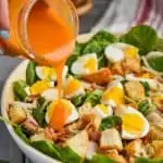 woman pouring red spinach salad dressing on a salad bowl filled with spinach, hard boiled eggs, and croutons