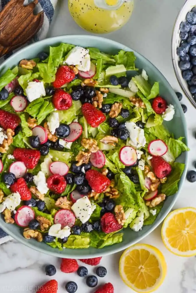 overhead view of a summer salad recipe with fresh raspberries, blueberries, radishes, romain lettuce, walnuts, and goat cheese all with a lemon poppyseed dressing in a big blue bowl