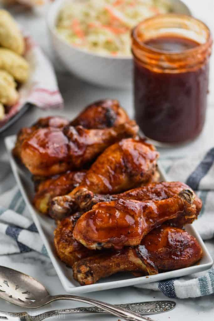 plate of grilled chicken legs on a rectangular white plate with a jar of bbq sauce and coleslaw in the background