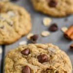 oatmeal chocolate chip cookie sitting on a blue wood board with chocolate chips and oats around it and two cinnamon sticks in the background