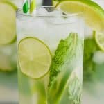 up close view of a high ball glass filled with a mojito cocktail, lime slices, fresh mint, striped green straws, and garnished with lime wedges and fresh mint
