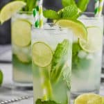 three high ball glasses on a white surface against a gray background filled with lime slices, a mojito recipe, fresh mint leaves, and garnished with green and white stripped straws, fresh mint and a lime wedge