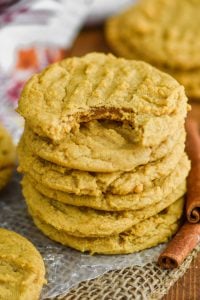 stack of pumpkin peanut butter cookies on wax paper on burlap with a cinnamon stick next to them and a bite out of the top cookie