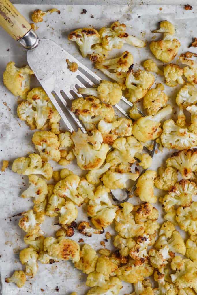overhead view of roasted cauliflower on a parchment lined baking sheet, being dished up by a slotted spatula