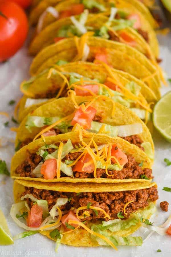 front view of a row of hard shell tacos with taco meet, and topped with tomatoes, lettuce, and cheese