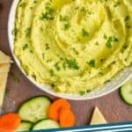 pinterest graphic of overhead view of a white bowl on a brown surface filled with edamame hummus recipe, garnished with parsley and oil and surrounded by cut vegetables and triangles of naan bread that says "edamame hummus simplejoy.com"