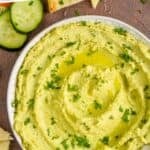 pinterest graphic of overhead view of a white bowl on a brown surface filled with edamame hummus recipe, garnished with parsley and oil and surrounded by cut vegetables and triangles of naan bread that says: "the best edamame hummus simplejoy.com"