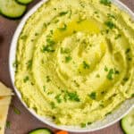 overhead view of a white bowl on a brown surface filled with edamame hummus recipe, garnished with parsley and oil and surrounded by cut vegetables and triangles of naan bread