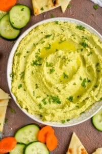 overhead view of a white bowl on a brown surface filled with edamame hummus recipe, garnished with parsley and oil and surrounded by cut vegetables and triangles of naan bread