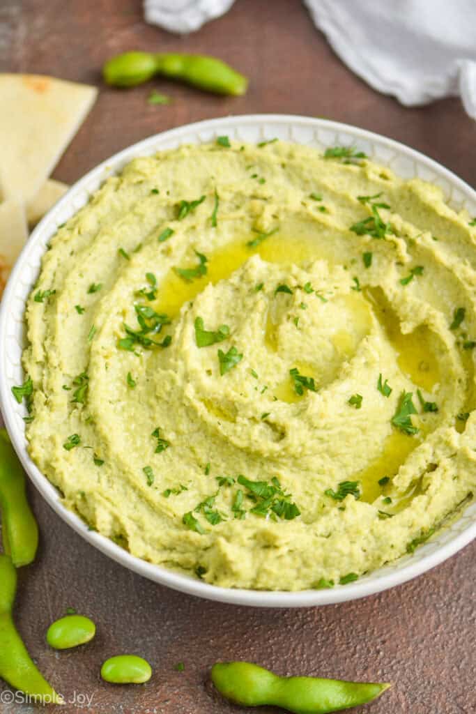 side view of edamame hummus recipe in a white bowl, garnished with parsley and oil with soy beans around the bowl