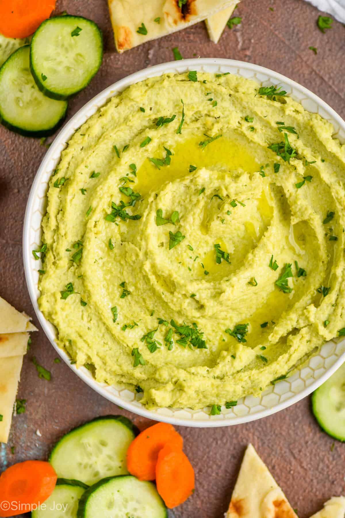 overhead view of a white bowl on a brown surface filled with edamame hummus recipe, garnished with parsley and oil and surrounded by cut vegetables and triangles of naan bread