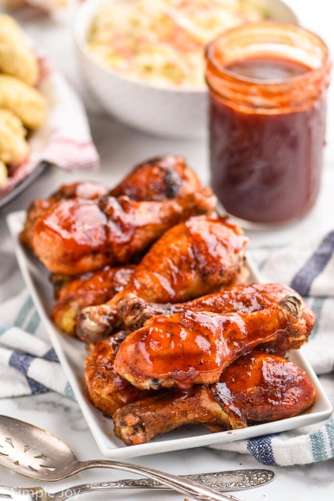 plate of grilled chicken legs on a rectangular white plate with a jar of bbq sauce and coleslaw in the background