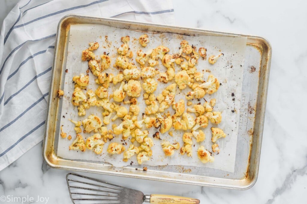 overhead view of roasted cauliflower recipe on a baking sheet that is lined with parchment paper, on a marble countertop with a striped kitchen towel underneath the baking sheet