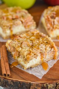 one apple crisp cheesecake bar on a square of wax paper, dripping with caramel sauce with two more bars in the background
