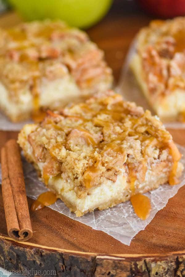 one apple crisp cheesecake bar on a square of wax paper, dripping with caramel sauce with two more bars in the background