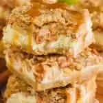 a stack of three apple cheesecake bars on wax paper on a wood platter with caramel sauce dripping from them and a bite out of the top one