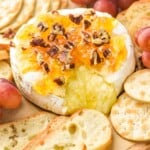 a wheel of baked brie with cheese coming out and topped with marmalade, honey, and pecans on a platter with baguette slices, crackers, and grapes - 8