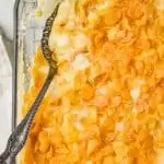 overhead view of a clear baking dish filled with cheesy potatoes with corn flakes with a spoon going into it