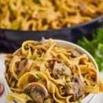 two stacked white bowls with the top one full of ground beef stroganoff recipe - egg noodles, mushrooms, parsley, and a skillet with the rest of the recipe in the background
