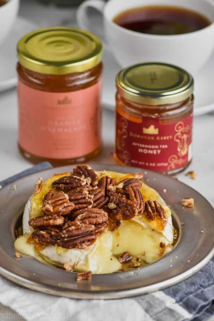 a silver plate with baked brie on it covered with honey, marmalade, pecans, and jars of honey and marmalade in the background