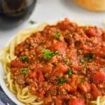white plate with spaghetti topped with spaghetti meat sauce recipe, two red wines and bread in the background, fork in the foreground
