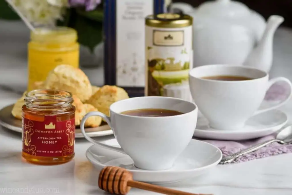 a tea cup next to a jar of honey, with a honey stick in front, another tea cup in back, as well as a canister of tea, a scone mix, and flowers