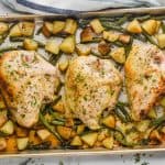 landscape photo of sheet pan chicken and vegetables with three bone in chicken breasts that have been cooked and nicely browned on a bed of cubed potatoes and green beans on a rimmed baking sheet on a white marble counter top with a striped blue kitchen towel and parsley pieces on the countertop
