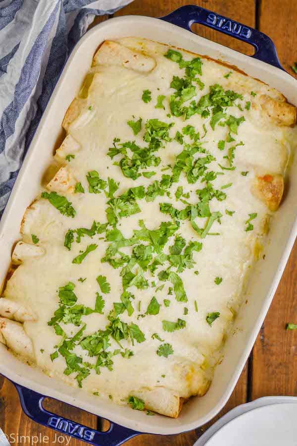 over head view of a baking dish with blue handles on a wood board holding sour cream chicken enchiladas, with a white sauce, garnished with a lot of cilantro