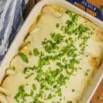 over head view of a baking dish with blue handles on a wood board holding sour cream chicken enchiladas, with a white sauce, garnished with a lot of cilantro