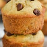 two banana chocolate chip muffins stacked on top of each other on top of wax paper with more muffins in the background