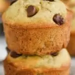 two banana chocolate chip muffins stacked on top of each other on top of wax paper with more muffins in the background