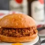 a homemade sloppy joe sitting on a metal and wood tray with chips around it and beer in the background