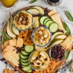 overhead view of a hummus charcuterie board with three different topped hummus bowls, cucumber slices, pieces of cheese, slices of pear, pretzel flats, and cranberries