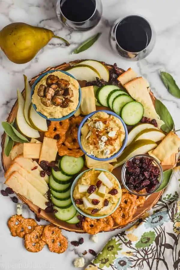 overhead view of a hummus charcuterie board with three different topped hummus bowls, cucumber slices, pieces of cheese, slices of pear, pretzel flats, and cranberries